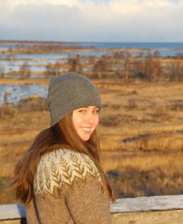 Young woman wearing a woolly sweater smiles at the camera. On the background, there is moraine ridge scenery which is typical for the Kvarken Archipelago.